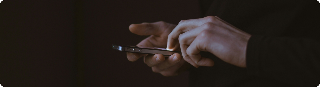 Hands holding a smartphone and tapping its glowing screen against a dark background.
