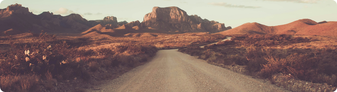 image alt text  A gravel road winds through Texas desert scrub towards rugged rock mountains in warm evening light.