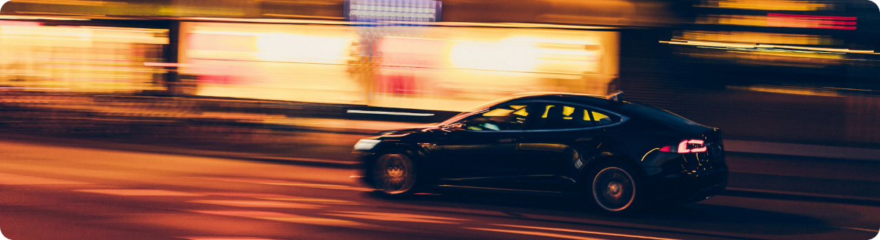 Black Tesla saloon speeding through a city at night, the background blurred into orange and yellow light.