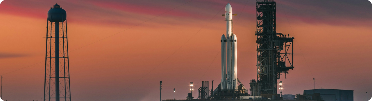 A SpaceX Falcon Heavy rocket on the launch pad beside its service tower at dusk, with a water tower against an orange sky.